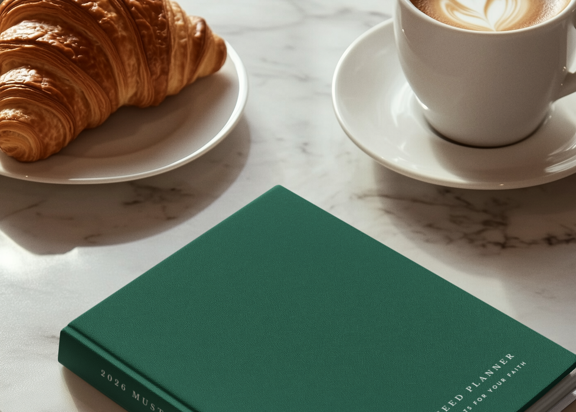 Green planner book on a marble table with croissants and a coffee cup.